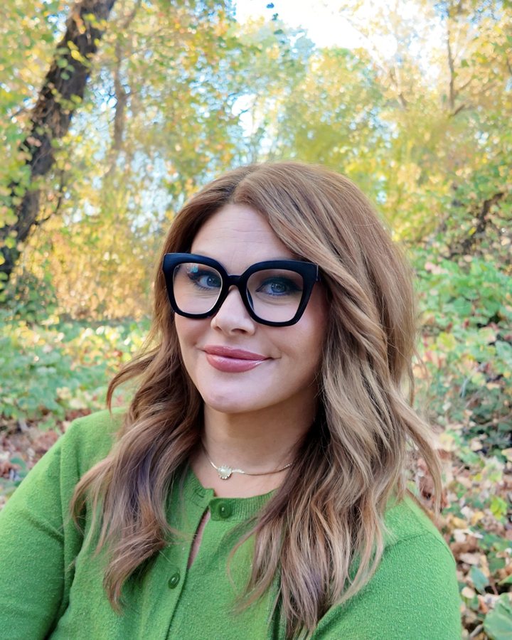 Dani, the founder of Bifocal, smiling in large black glasses and a green cardigan, outdoors among autumn leaves.